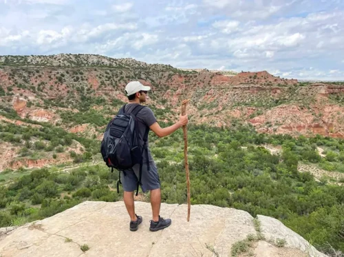 A hiker with a backpack and walking stick stands on a rocky ledge overlooking Palo Duro Canyon State Park.