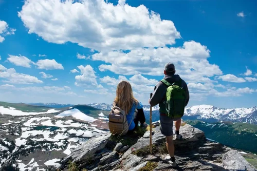 Two hikers with backpacks admire the snow-capped mountains and blue sky in the Rocky Mountains.