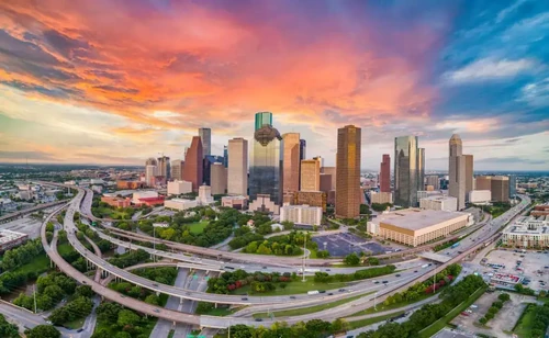A sunset behind downtown Houston, Texas, with skyscrapers, highways, and lush green spaces.