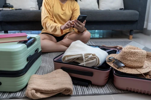 A young woman sits on the floor packing a suitcase while using her phone to plan her first road trip.