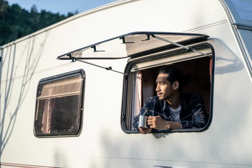 A young man drinking coffee while looking out his RV window.