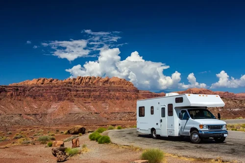 A white RV parked on a scenic desert road with red rock formations and a bright blue sky with clouds in the background.